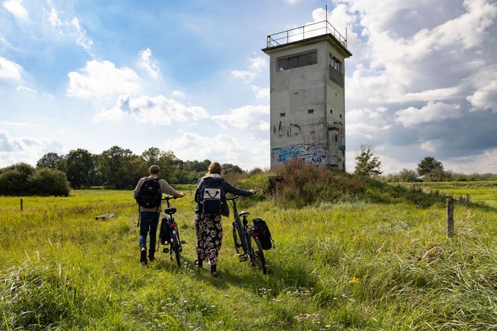Eine Wiesenlandschaft mit einem rechteckigen Turm, der mit Graffity besprüht ist. Eine Frau und ein Mann schieben jeweils ein Fahrrad